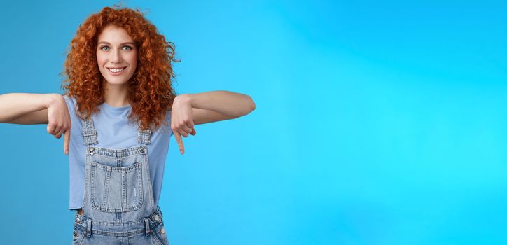 Lifestyle. Charismatic sassy flirty redhead daring ginger girl curly haircut pointing down index fingers smiling broadly enthusiastic explore new store pointing promo like cool advertisement blue background.