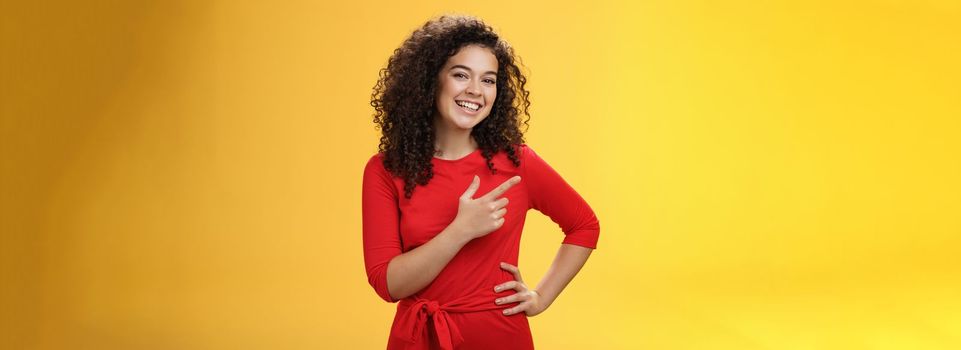 Portrait of friendly and assertive charming female coworker showing around newbie pointing left and smiling, laughing at camera having fun posing with hand on hip over yellow background. Advertisement concept