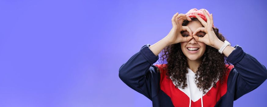 Girl see us as imitating peek with binocular making circles over eyes and looking through it joyful and excited, having fun fooling around posing happy and cute over blue background in outdoor outfit.