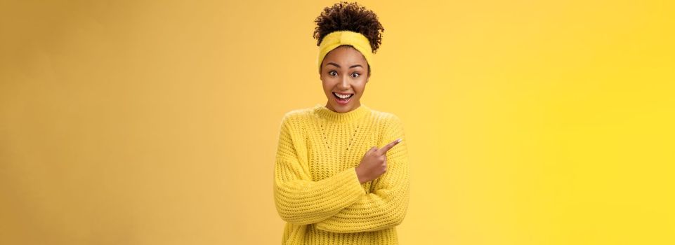 Amused charismatic smiling black cute girl. in headband sweater widen eyes drop jaw astonished hear about awesome interesting new place talking friend standing yellow background pointing left amazed.