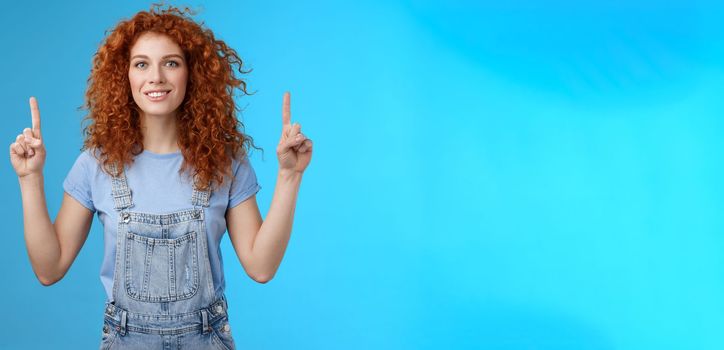 Motivated happy cheerful redhead silly curly woman pointing up inrdex finger smiling charmed impressed excited showing awesome promo discuss interesting advertisement blue background.