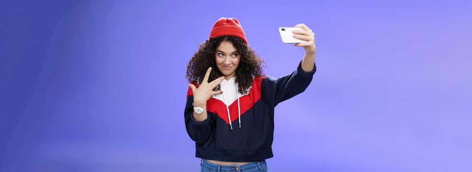 Lifestyle. Cool and stylish good-looking caucasian female with curly hair in trendy red beanie and sweatshirt looking from under forehead awesome and swah taking selfie with smartphone holded in extended arm.