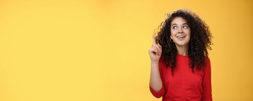 I got idea. Excited happy attractive woman with curly hair in red dress raising index finger in eureka gesture smiling amused, looking at upper left corner, sharing plan or suggestion over yellow wall.