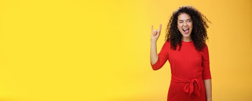 Rebellious meloman leaving in tender girl. Thrilled and carefree curly-haired woman in red dress sticking out tongue and looking right with smile as showing rock-n-roll gesture, enjoying cool music.