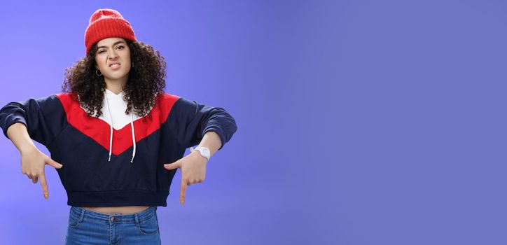 Lifestyle. Snobbish and displeased stylish and cool street girl with curly hair in beanie and sweatshirt pointing down grimacing as feeling unsatisfied and disappointed posing against blue background.