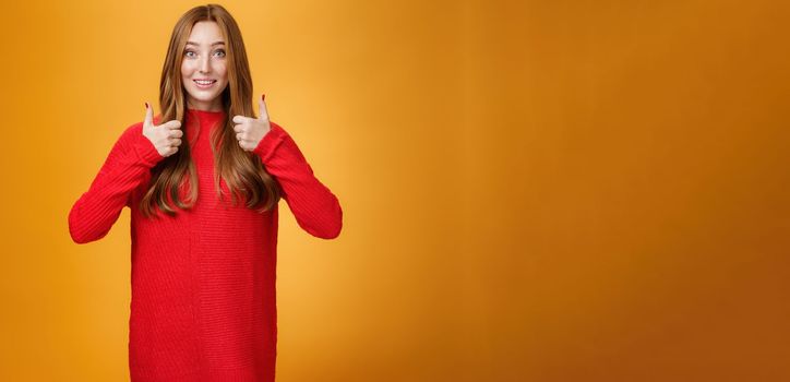 Excited and supportive redhead girlfriend asking opinion about new warm dress showing thumbs up and raising eyebrows questioned as waiting for opinion over orange background.