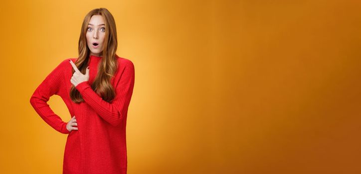 Lifestyle. Impressed redhead girl reacting to awesome unbeliavable promotion pointing behind or at upper left corner folding lips staring at camera astonished and surprised over orange background.