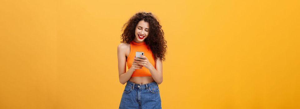 Girl spending time in internet texting friend messages. via smartphone laughing while looking at device screen standing happy and upbeat over orange background in cropped top and denim shorts.