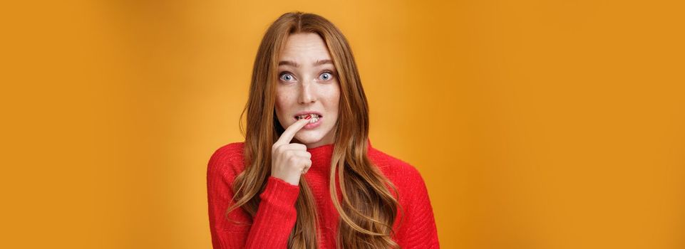 Silly, cute redhead woman making innocent oops expression holding finger on lip and looking scared with questioned expression at camera making mistake and trying get away of troubles with flirty gaze.