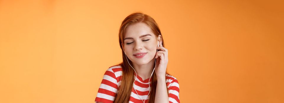 Lovely attractive redhead caucasian 20s woman listen music, close eyes tender smile, touch earphone ear, tilt head enjoyment and delight, hear nice beats, favorite music track, orange background.