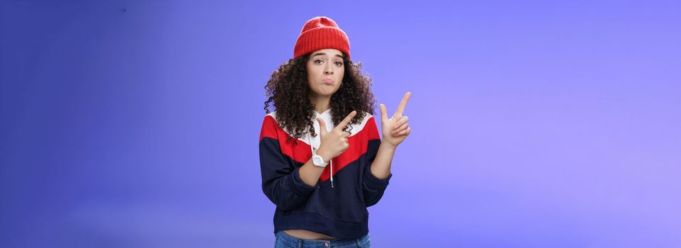 Portrait of silly upset cute curly-haired woman in trendy beanie pursing lips in sadness and frowning making disappointed, regret face pointing at upper left corner unhappy and sad over blue wall.