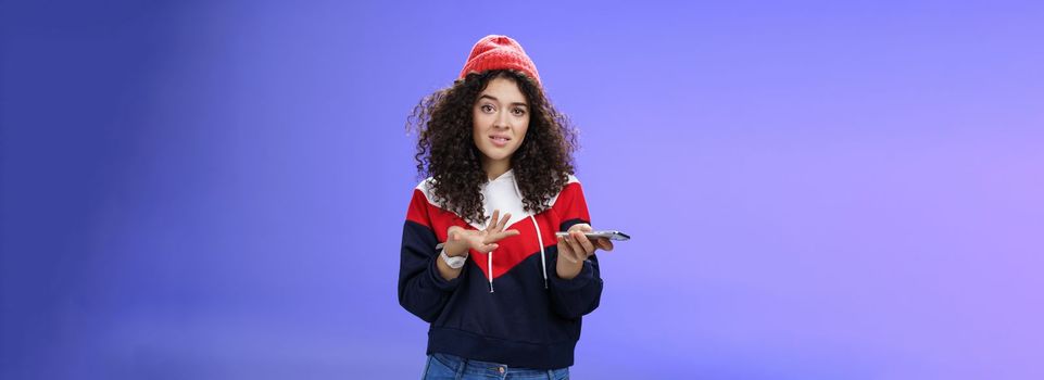 Confused and clueless girl cannot understand meaning of strange message shrugging grimacing questioned as holding smartphone and extending arm in unaware gesture over blue background.