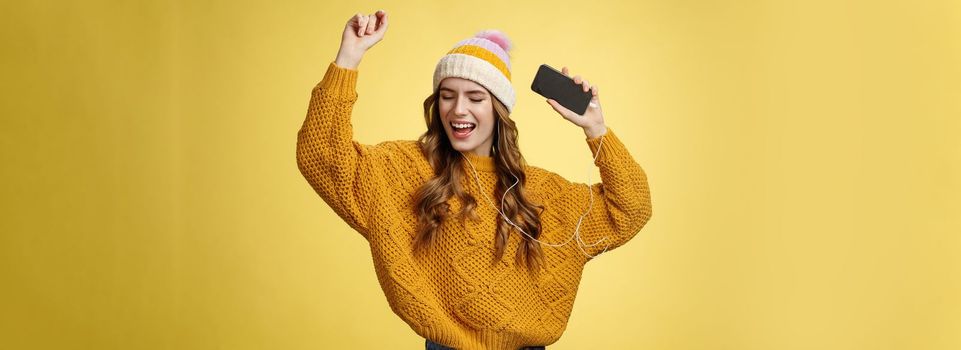 Carefree happy girl enjoying listen music wearing wired earphones raising hands dancing joyfully having fun singing along awesome song playing playlist holding smartphone, standing yellow background.