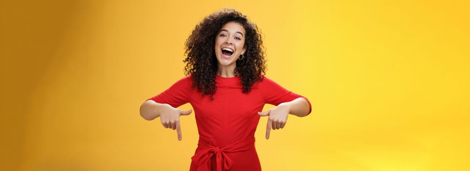 Look what I got here. Charismatic carefree happy charming woman with curly hair in red dress laughing with broad smile pointing down as showing awesome copy space to customers over yellow wall.