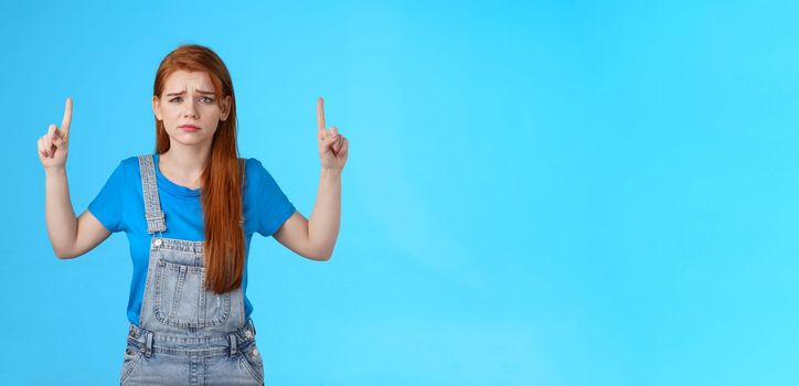 Doubtful concerned unsure redhead female making important decision, grimacing hesitant displeased, point up promo, top copy space, feel uncertain unlikely buy suspicious product, blue background.