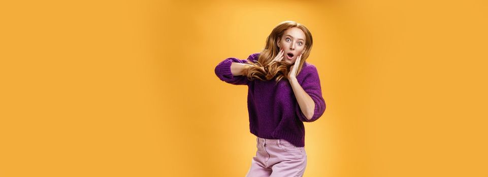 Lifestyle. Impressed and surprised amazed good-looking redhead female in purple sweater open mouth astonished and holding hands on cheeks as jumping popping eyes amused over orange background.