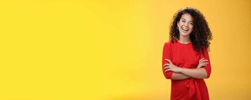 Self-assured happy enthusiastic curly-haired female reporter in cute red dress laughing carefree, having fun tilting head amused and holding hands crossed over body in confident pose over yellow wall.