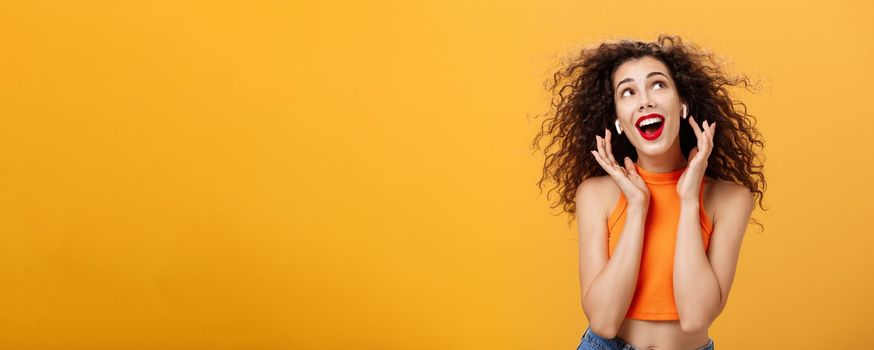 Waist-up shot of attractive silly curly-haired european female. in cropped top using wireless earphones touching earbuds and gazing at upper right corner delighted and carefree over orange wall.