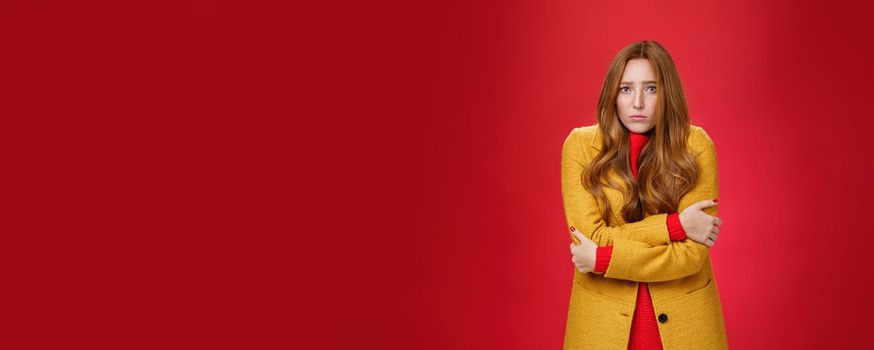 Cute and sad trembling redhead girl in light yellow coat shaking from cold, having goosebumps frowning and pouting from sorrow as freezing on windy weather over red background. Copy space