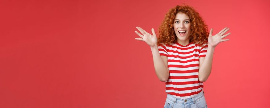 Awesome incredible performance bravo. Attractive cheerful cute european redhead curly female raise hands thrilled express excitement happiness joy look camera entertained delighted red background.