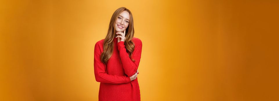 Sweet and tender redhead woman with cute freckles in red knitted dress tilting head touching face with fingers and smiling delighted and loving a camera over orange background. Copy space