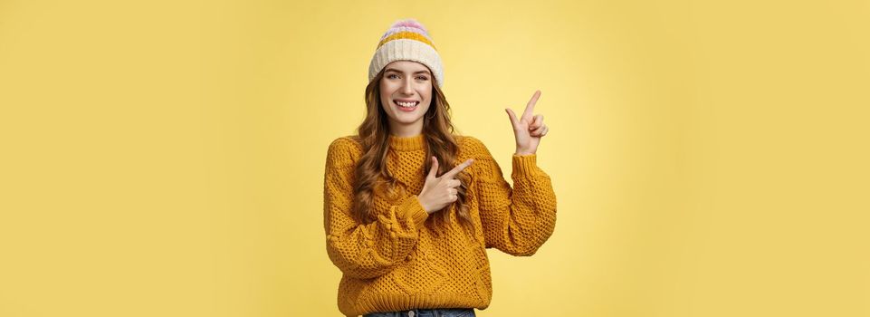 Charming carefree smiling young 20s woman wearing hat sweater pointing upper left corner sideways sharing interesting link awesome promotion delighted recommend cool product, yellow background.