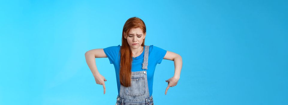 Moody upset cute redhead woman feel uneasy, frowning look pointing up jealousy and regret, cannot afford awesome new product, express sadness and sorrow, stand blue background gloomy.