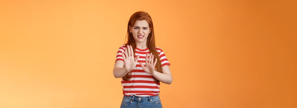 Gosh it stinks. Disgusted redhead picky woman blocking sign raise hands up defensive, grimacing, cringe from aversion awful smell, show refusal rejecting disgusting offer, stand orange background.