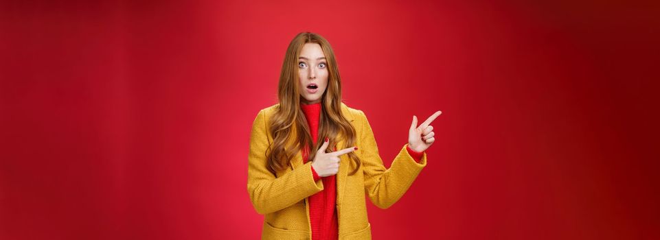 Stunned speechless and impressed attractive redhead woman with freckles in yellow coat drop jaw amazed as popping eyes at camera questioned, pointing left astonished over red background.