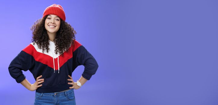 Lifestyle. Confident ad energized cute european woman with curly hairstyle in warm beanie and stylish sweatshirt holding hands on waist and smiling friendly at camera as ready to go out and have fun outside.