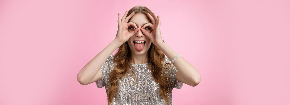 Carefree playful funny young party girl fool around showing tongue immature mimicking make goggles from hands widen eyes look through circles, having fun entertain friends, standing pink background.