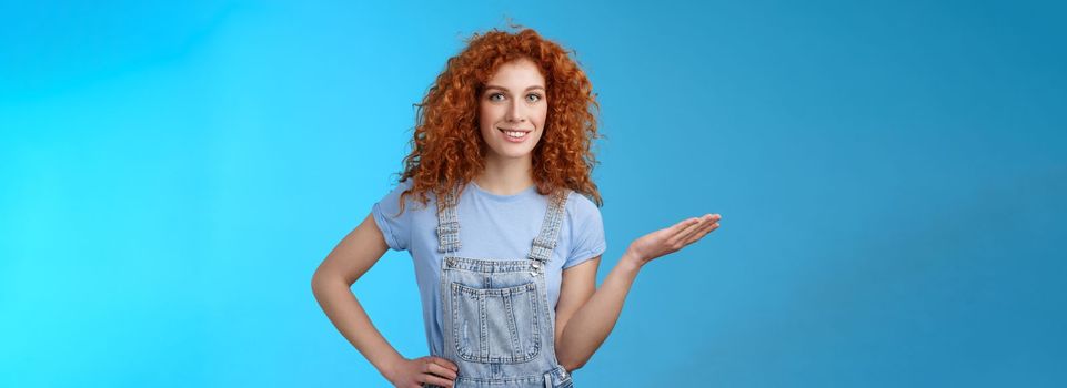 Let me show you cool product. Attractive cheerful confident redhead female model curly hairstyle present customers object hold palm blank blue copy space advertising standing blue background.
