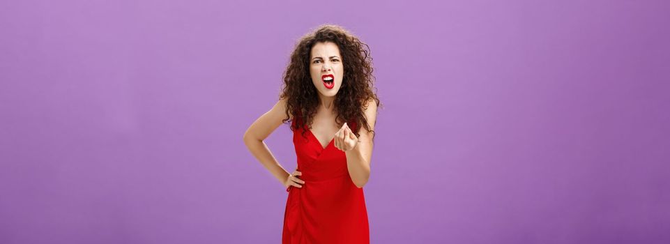 Woman in fury fighting with husband over money. making italian gesture with fingers shouting and yelling from fury and anger standing displeased and pissed over purple wall in red elegant dress.