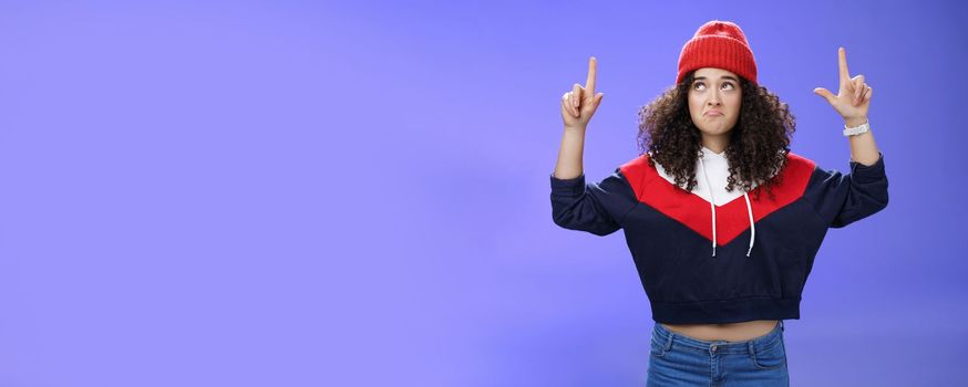 Girl feeling disappointed and gloomy as looking at bad weather making sad smile, pursing lips down raising hands staring up upset and unsatisfied standing displeased against blue background.