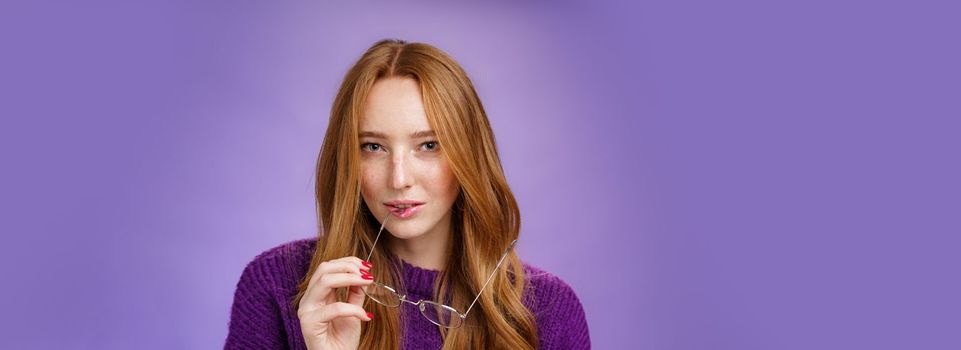 Close-up shot of smart and creative female redhead genius biting frame of glasses and squinting curious at camera as thinking having idea, making up plan against purple background.