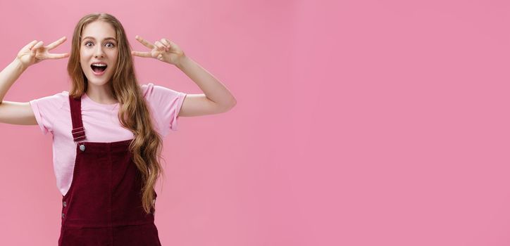 Girl staying positive lifting after falling. Charming carefree friendly-looking young woman with small scar on arm and tattoo showing peace gestures, open mouth and looking at camera over pink wall. Body language, body-positive, skinpositivity concept