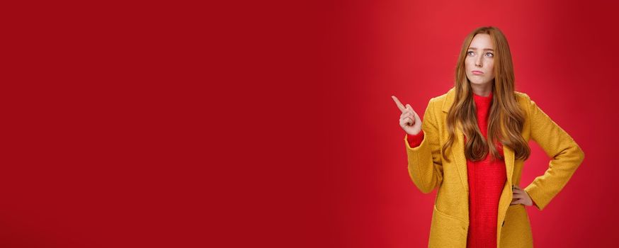 Sad and gloomy cute ginger girl in 20s wearing trendy yellow coat holding hand on waist, pouting, looking and pointing at upper left corner with regret and sorrow over red background.