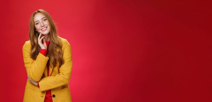 Good-looking carefree and happy relaxed redhead girlfriend in yellow stylish coat touching face and tilting head as smiling with positive emotions, posing cheerful against red background.