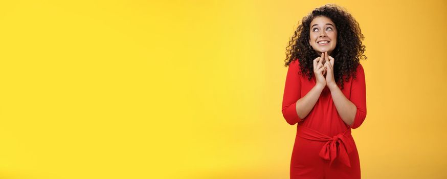 Hopeful, excited cute tender woman in red dress with curly hair standing new x-mass tree crossing fingers for good luck and smiling with head raised to sky making wish, having faith in dream come true.