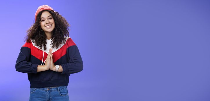 Portrait of charming curly-haired feminine girlfriend begging for favour as posing over blue background in winter outfit with hat, holding hands in pray smiling with angel look and friendly gaze. Body language and facial expressions concept