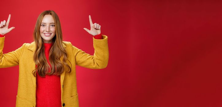 Indoor shot of redhead attractive woman in yellow fall coat raising hands promoting advertisement as pointing up and smiling broadly with satisfied pleasant expression over red background. Copy space