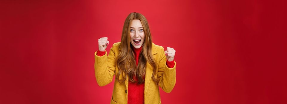 Studio shto of cheerful excited good-looking ginger girl in yellow coat raising clenched fists in joy and happiness, triumphing yelling yes from success and triumph, celebrating win over red wall.