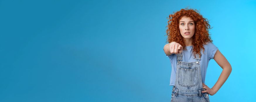 You seem familiar. Worried concerned lively redhead emotive curly girl pointing camera look excited nervously recognize someone standing confused hold hand waist wear overalls. Copy space