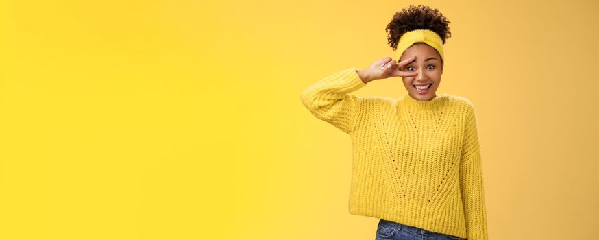 Silly insecure cute awkward young millennial girl blushing unconfident photographing smiling show peace victory sign near eye bad in posing, standing friendly yellow background. Copy space