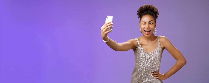 Stylish sassy african american b-day girl taking selfie new elegant silver shiny dress extend arm holding smartphone posing winking display screen amused having fun smiling broadly, blue background.