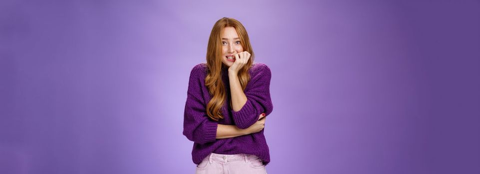 Lifestyle. Portrait of nervous insecure cute redhead girl in purple sweater biting fingernails and stooping as feeling scared before exam or interview standing anxious and unsure over violet background.