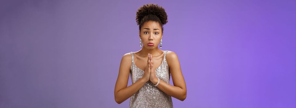 Attractive african-american woman in glittering silver evening dress begging friend favour look dearly pouting frowning silly face asking please give me money lend outfit, blue background.