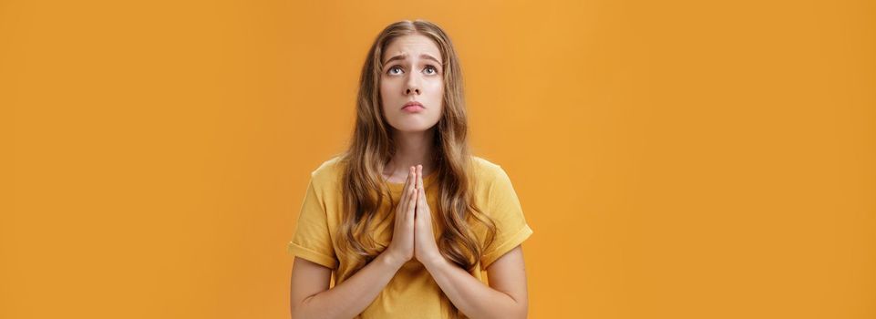 Hopeful uneasy and worried gloomy faithful girl in t-shirt holding hands in pray against chest looking up with sad look praying making wish for good well of family posing over orange wall. Body language concept