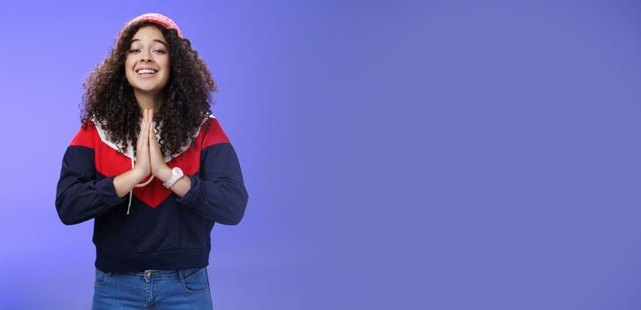 Lifestyle. Charming excited and friendly-looking cute female friend with curly hairstyle in outdoor hat and sweatshirt holding hands in pray and smiling with angel expression as hoping friend help over blue wall.