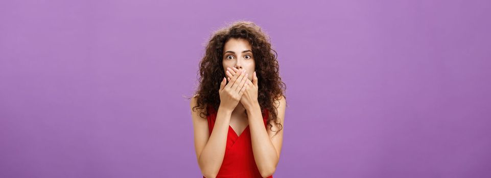Cocnerned and shocked troubled adult woman with curly hairstyle in elegant evening red dress gasping from surprise and fear covering mouth with palms standing speechless over purple wall. Copy space